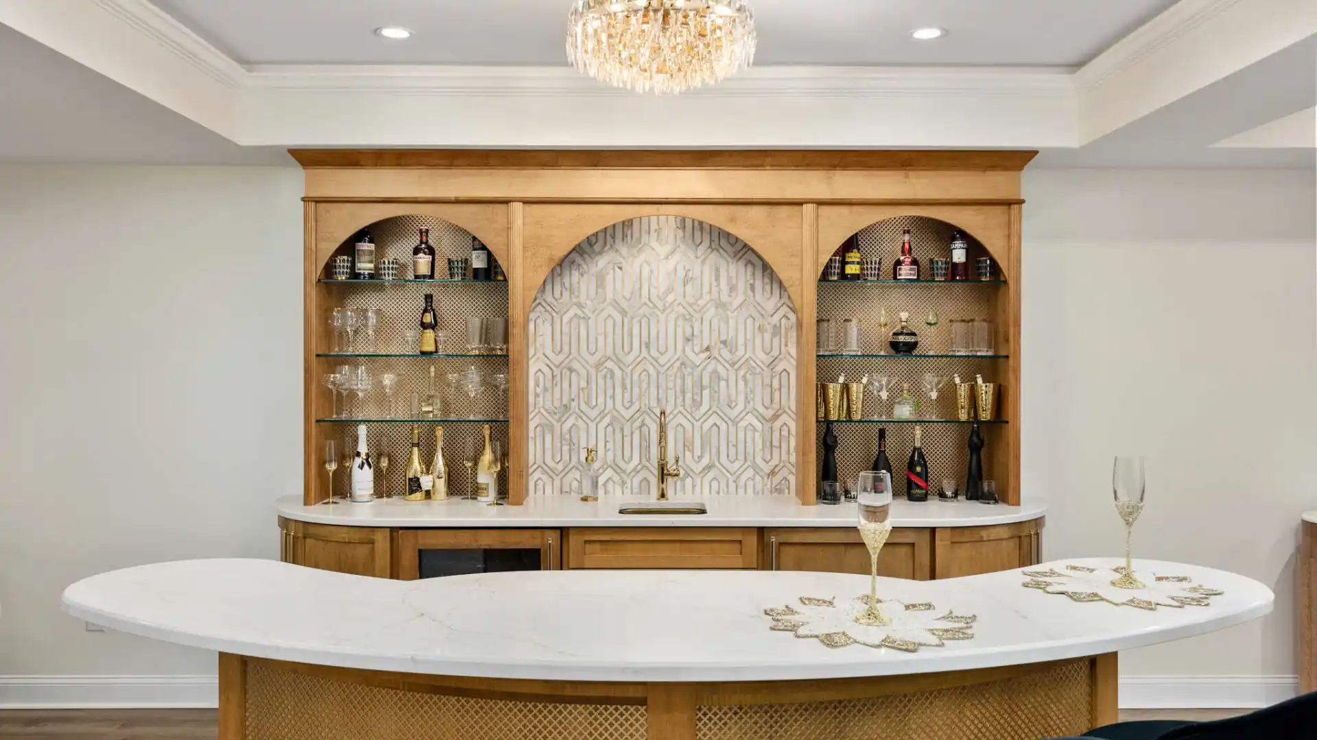 A stylish home bar with a curved white countertop and gold accents. Wooden shelves with decorative bottles flank an ornate patterned backsplash. A crystal chandelier hangs above, illuminating the space elegantly—perfect for discussing outdoor living in Delaware County, PA over wine.