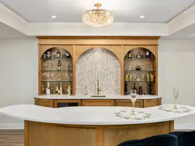 A stylish home bar with a curved white countertop and gold accents. Wooden shelves with decorative bottles flank an ornate patterned backsplash. A crystal chandelier hangs above, illuminating the space elegantly—perfect for discussing outdoor living in Delaware County, PA over wine.