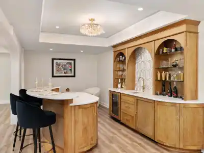 A modern home bar with wooden cabinets, a marble countertop, and a curved bar area overlooking exquisite outdoor living. Various bottles and glasses are on display. Three black bar stools stand in front, while a decorative chandelier hangs from the ceiling, perfectly complementing the Delaware County vibe.