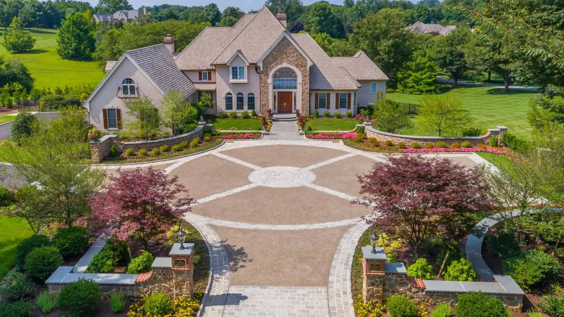 Aerial view of a large, elegant house with a circular driveway and landscaped gardens. The home features a beige exterior with multiple gables, a stone chimney, and a grand entrance. Enhance your outdoor living in Delaware County PA with professional landscaping services for lush greenery and vibrant flowerbeds.