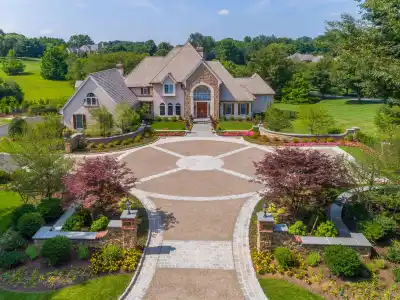 Aerial view of a large, elegant house with a circular driveway and landscaped gardens. The home features a beige exterior with multiple gables, a stone chimney, and a grand entrance. Enhance your outdoor living in Delaware County PA with professional landscaping services for lush greenery and vibrant flowerbeds.