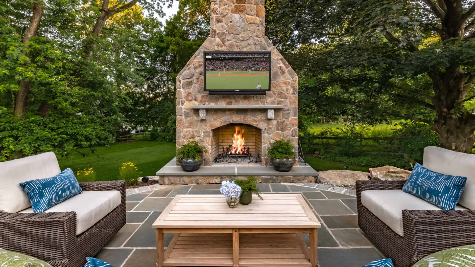 Outdoor living in Delaware County, PA, shines on this patio with wicker couches adorned with blue and white cushions, a wooden coffee table with flowers, and a large stone fireplace. A flat-screen TV shows a sports match as lush greenery envelops the space.