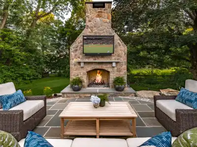 Outdoor living in Delaware County, PA, shines on this patio with wicker couches adorned with blue and white cushions, a wooden coffee table with flowers, and a large stone fireplace. A flat-screen TV shows a sports match as lush greenery envelops the space.