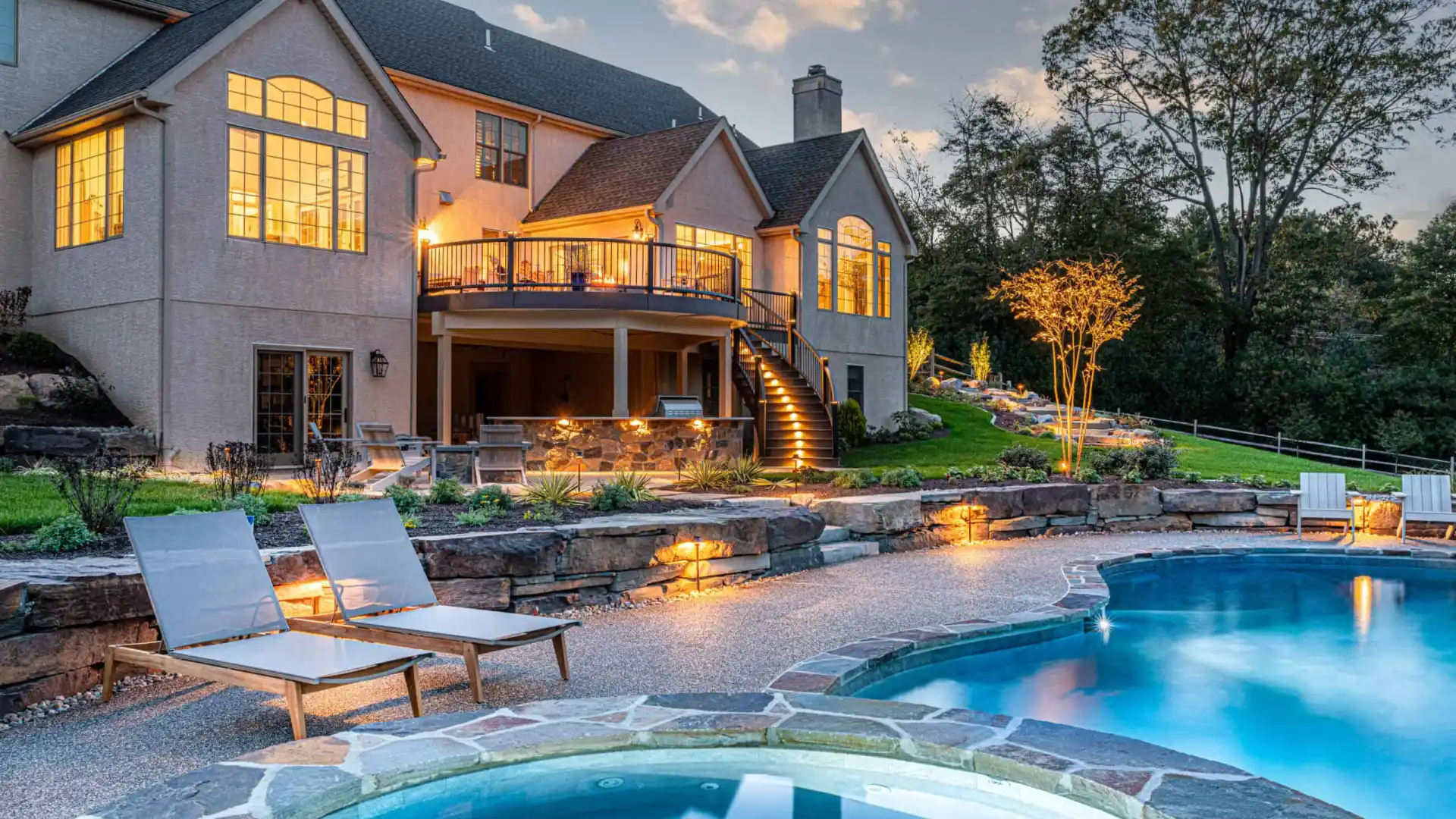 Luxurious house with large windows and a spacious deck, overlooking a well-lit backyard at dusk. Features a swimming pool with a hot tub in the foreground and two lounge chairs nearby, surrounded by expertly crafted landscaping services in Delaware County PA.
