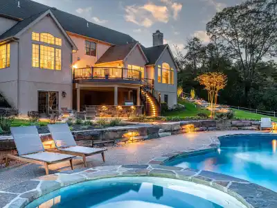 Luxurious house with large windows and a spacious deck, overlooking a well-lit backyard at dusk. Features a swimming pool with a hot tub in the foreground and two lounge chairs nearby, surrounded by expertly crafted landscaping services in Delaware County PA.