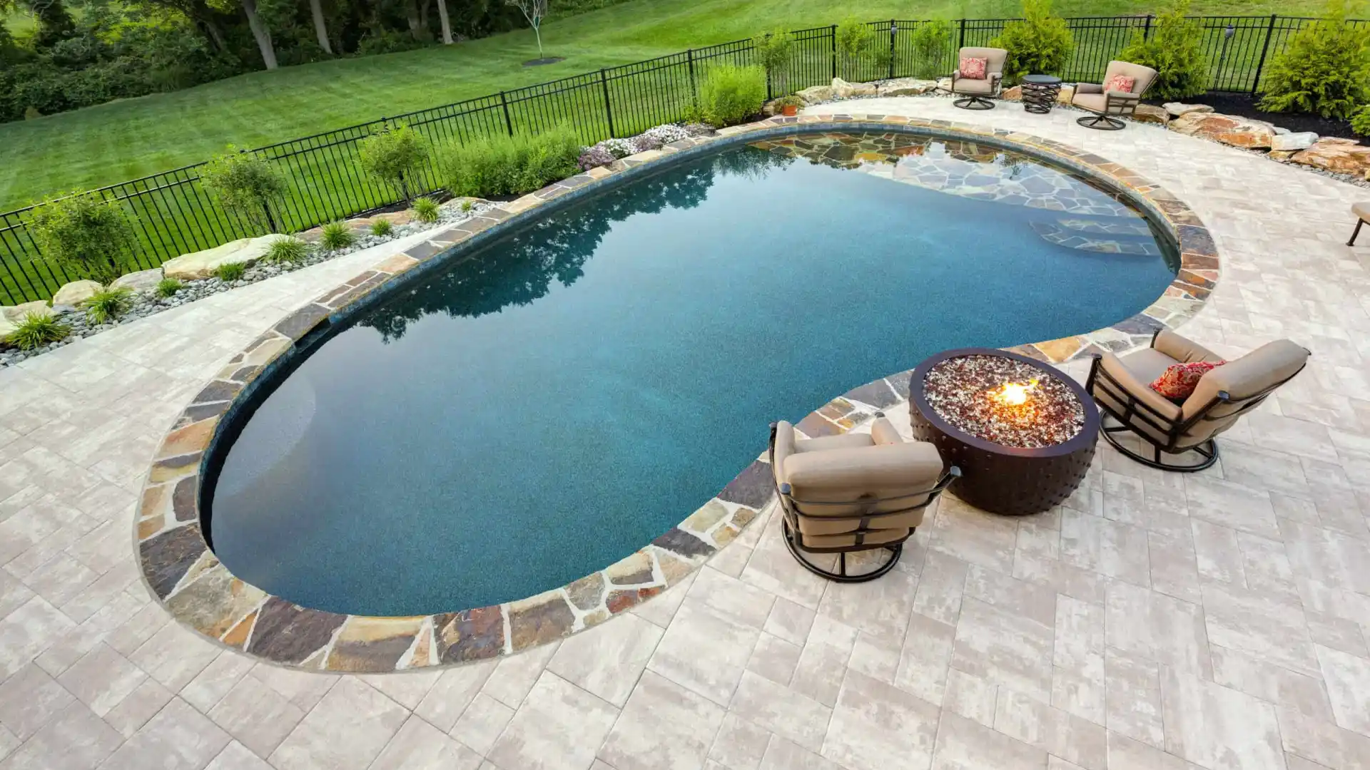 A backyard in Delaware County, PA features an oval swimming pool bordered by stones and surrounded by a paved patio. Two cushioned chairs face a circular fire pit, while lush grass and trees enhance the serene atmosphere. Perfectly crafted with expert hardscaping services to elevate outdoor living.