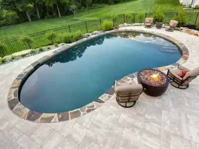 A backyard in Delaware County, PA features an oval swimming pool bordered by stones and surrounded by a paved patio. Two cushioned chairs face a circular fire pit, while lush grass and trees enhance the serene atmosphere. Perfectly crafted with expert hardscaping services to elevate outdoor living.