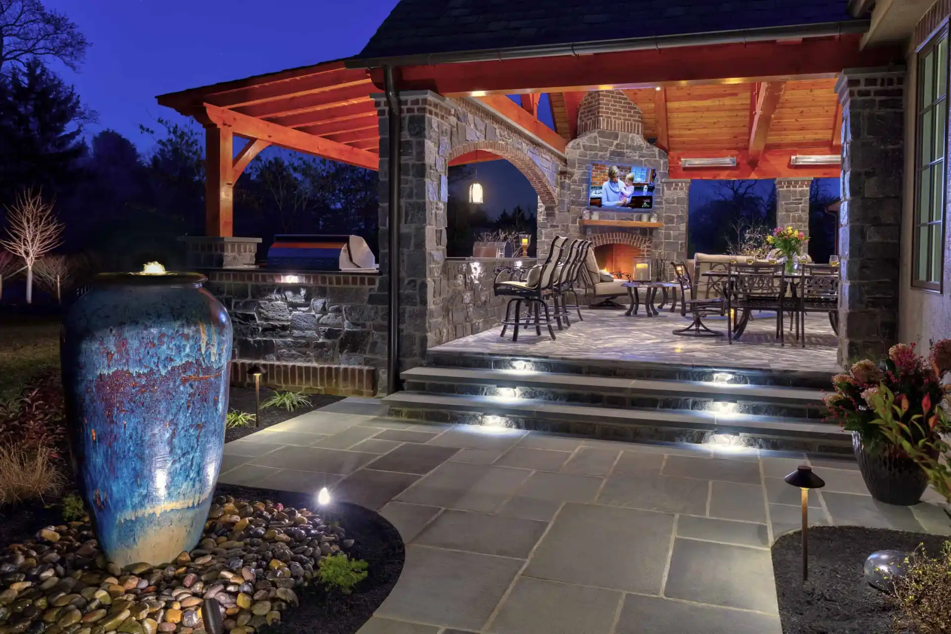 This cozy outdoor pavilion at night, crafted from stone and wood, boasts a glowing fireplace and inviting seating area. A large blue ceramic vase highlights the space, with subtle lighting enhancing the stone walkway and landscaped charm, epitomizing outdoor living in Delaware County, PA.