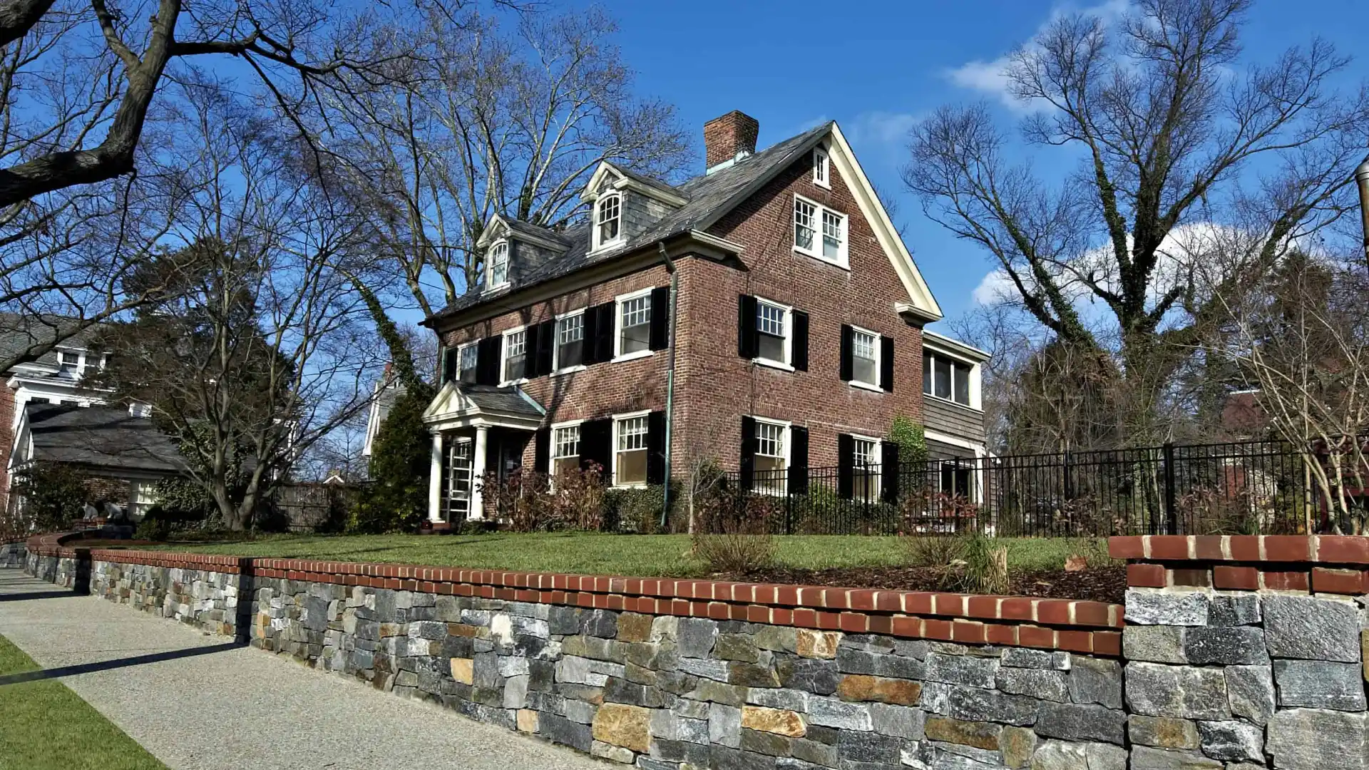 A large brick house with white trim sits behind a low stone wall and a sidewalk, enhanced by professional landscaping services in Delaware County, PA. It features multiple windows, a prominent chimney, and a gabled roof. Leafless trees surround the property under a clear blue sky.