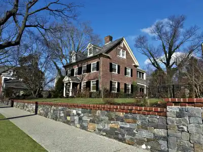 A large brick house with white trim sits behind a low stone wall and a sidewalk, enhanced by professional landscaping services in Delaware County, PA. It features multiple windows, a prominent chimney, and a gabled roof. Leafless trees surround the property under a clear blue sky.