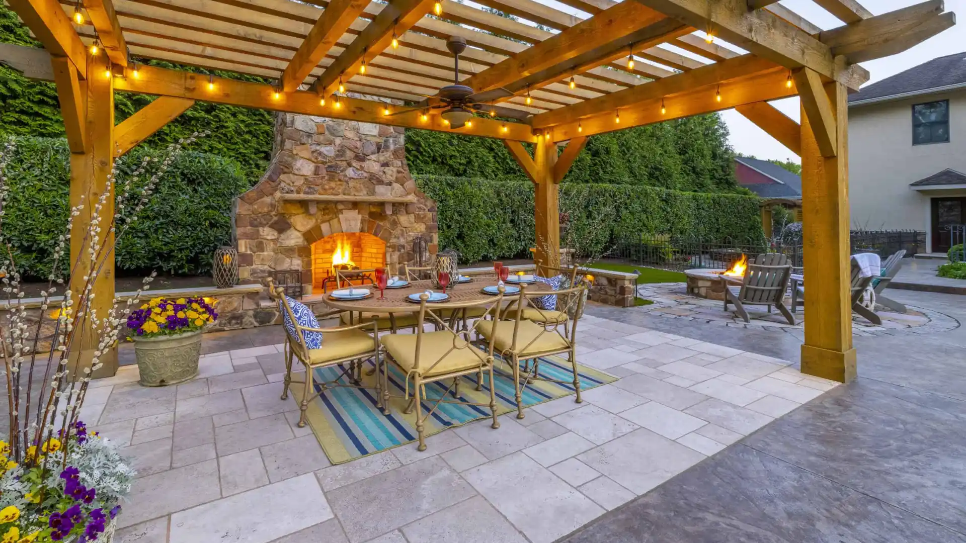 Outdoor living in Delaware County, PA comes alive with a wooden pergola adorned with string lights. A lit stone fireplace and a fire pit add warmth, while the dining table set on a tiled floor with rugs, potted plants, and comfy chairs enhance this beautifully hardscaped scene.