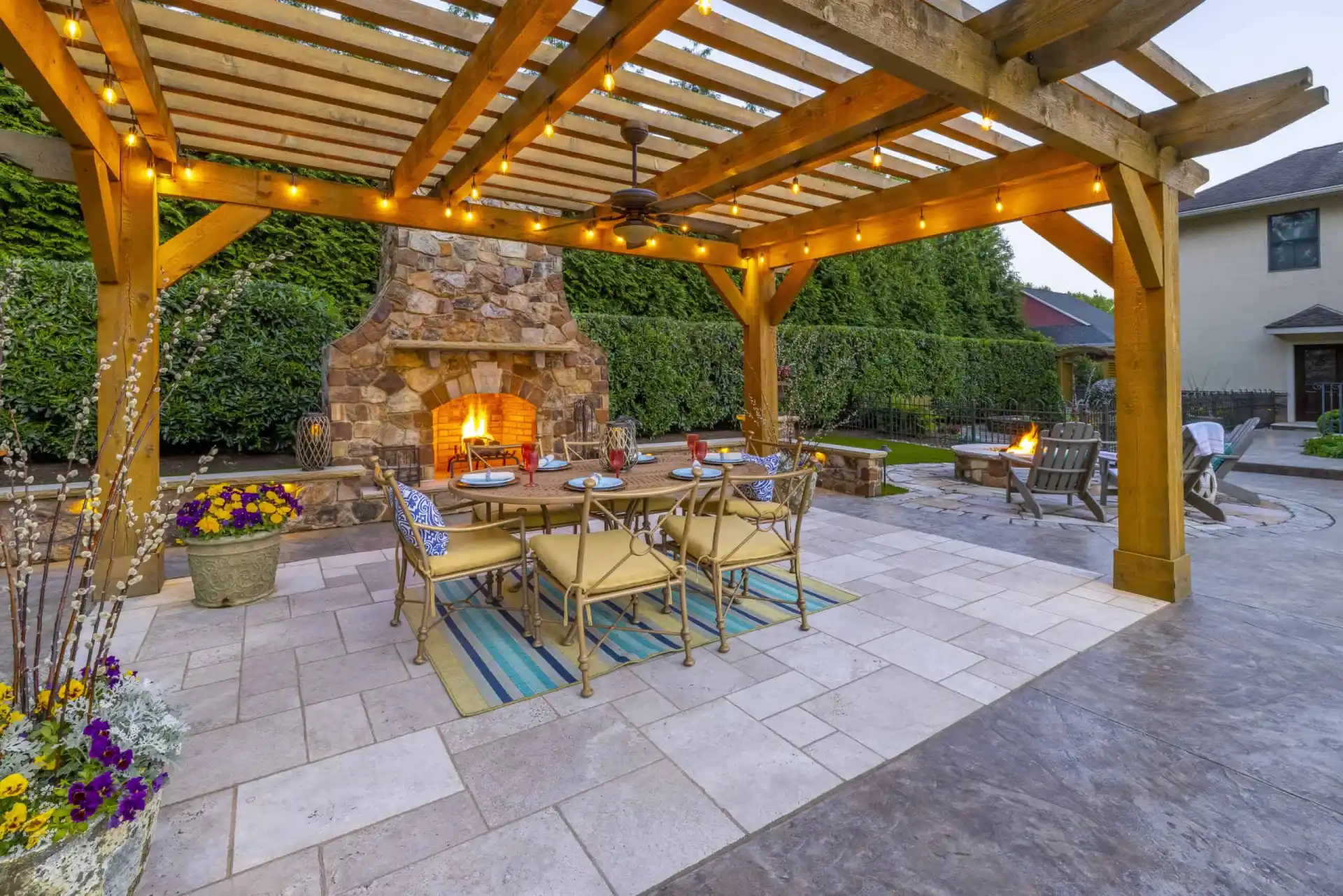 Outdoor living in Delaware County, PA comes alive with a wooden pergola adorned with string lights. A lit stone fireplace and a fire pit add warmth, while the dining table set on a tiled floor with rugs, potted plants, and comfy chairs enhance this beautifully hardscaped scene.