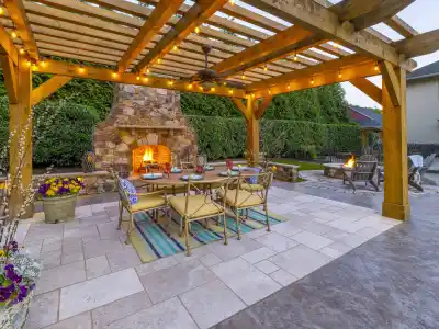 Outdoor living in Delaware County, PA comes alive with a wooden pergola adorned with string lights. A lit stone fireplace and a fire pit add warmth, while the dining table set on a tiled floor with rugs, potted plants, and comfy chairs enhance this beautifully hardscaped scene.
