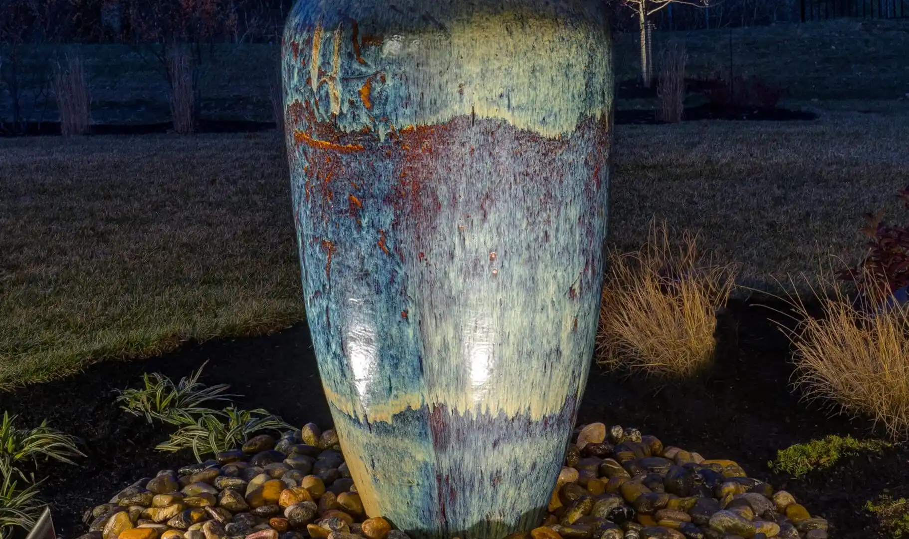 A tall, illuminated ceramic fountain vase sits on a bed of pebbles in a landscaped garden at night, showcasing the artistry of outdoor living in Delaware County, PA. The vase features a textured, multicolored glaze amidst trees and grass under a dark, clear sky.
