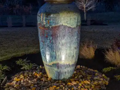 A tall, illuminated ceramic fountain vase sits on a bed of pebbles in a landscaped garden at night, showcasing the artistry of outdoor living in Delaware County, PA. The vase features a textured, multicolored glaze amidst trees and grass under a dark, clear sky.