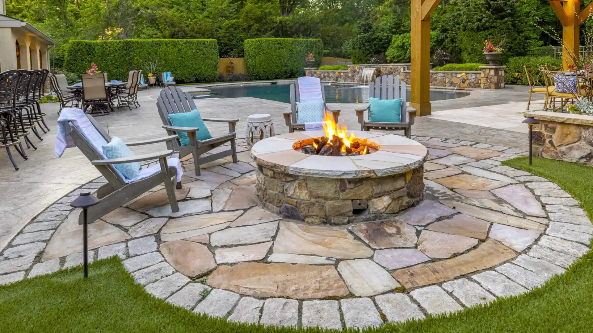 A landscaped backyard in Delaware County, PA, showcases outdoor living at its finest with a stone fire pit and wooden chairs with blue cushions. Behind it, a pool and patio furniture rest beneath a pergola adorned with string lights, all framed by lush greenery.