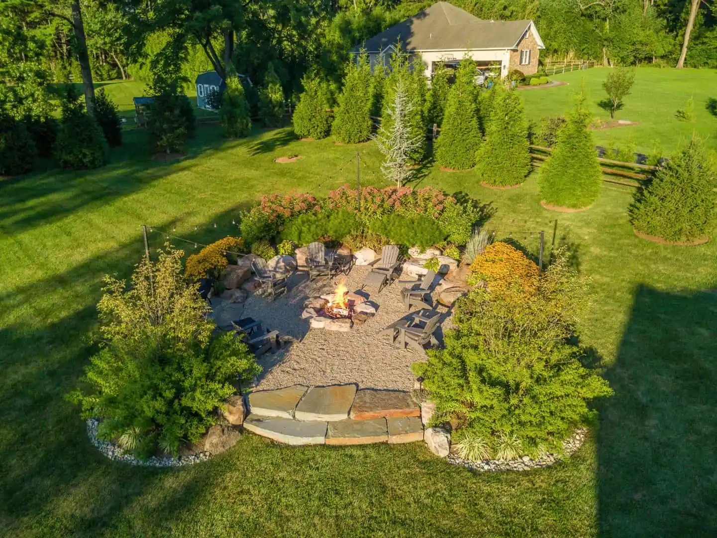 Aerial view of a circular stone patio with a fire pit, crafted expertly by hardscaping services in Delaware County PA. Surrounded by chairs, lush greenery, and trees encircle the patio. A house is visible in the background set within a large, grassy area.