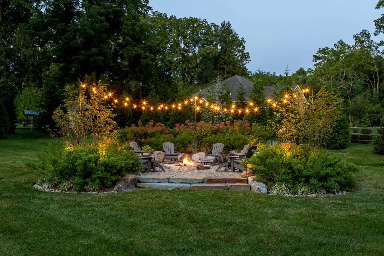 A circular outdoor fire pit area with Adirondack chairs amidst lush greenery and trees showcases exquisite outdoor living in Delaware County, PA. String lights hang above, enhancing the cozy atmosphere. In the background, a house peeks through the trees.