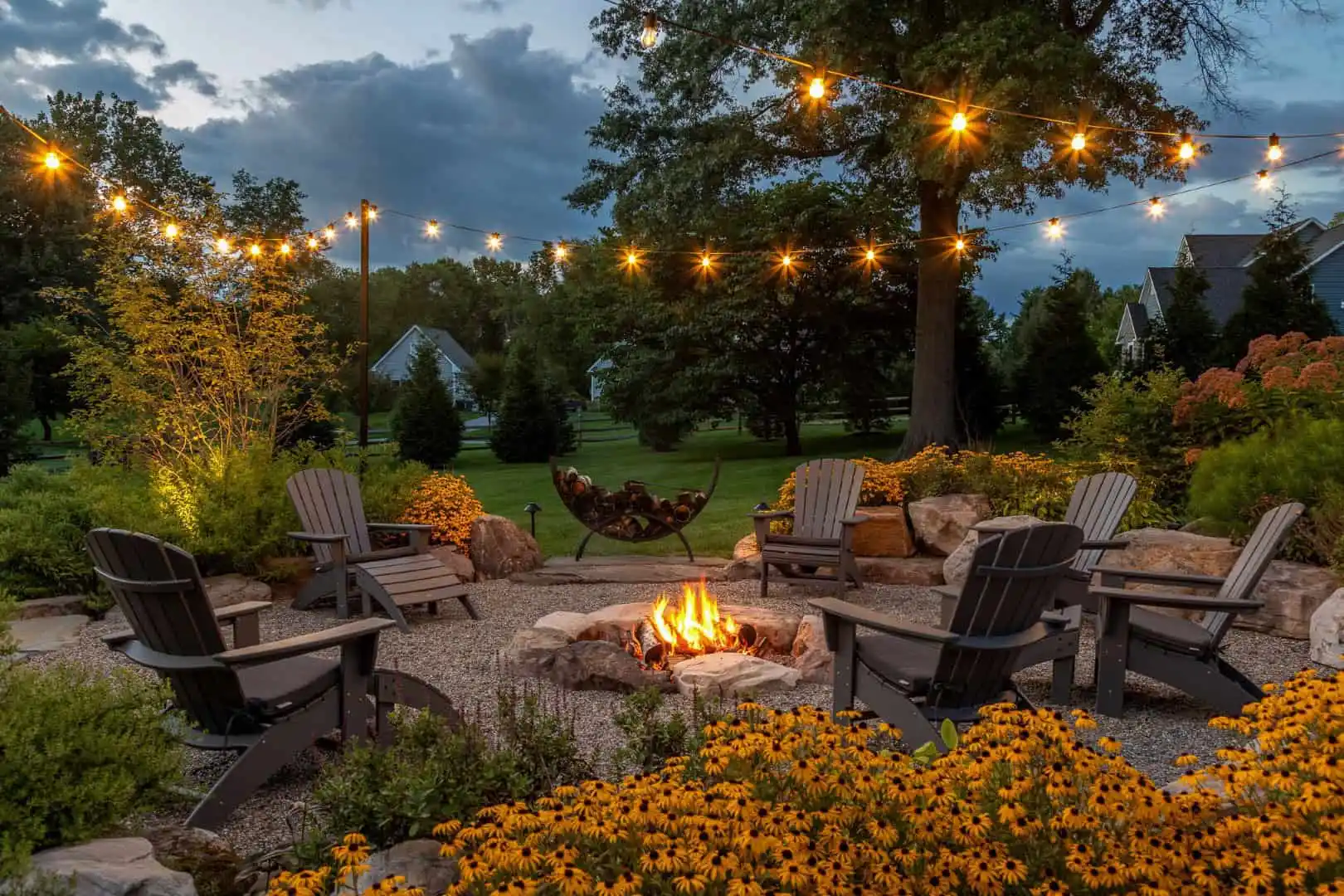 A cozy outdoor living scene in Delaware County, PA, features five Adirondack chairs around a fire pit, embraced by lush greenery and yellow flowers. String lights dangle overhead while houses and trees fade into the dusk backdrop.