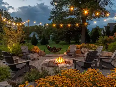 A cozy outdoor living scene in Delaware County, PA, features five Adirondack chairs around a fire pit, embraced by lush greenery and yellow flowers. String lights dangle overhead while houses and trees fade into the dusk backdrop.