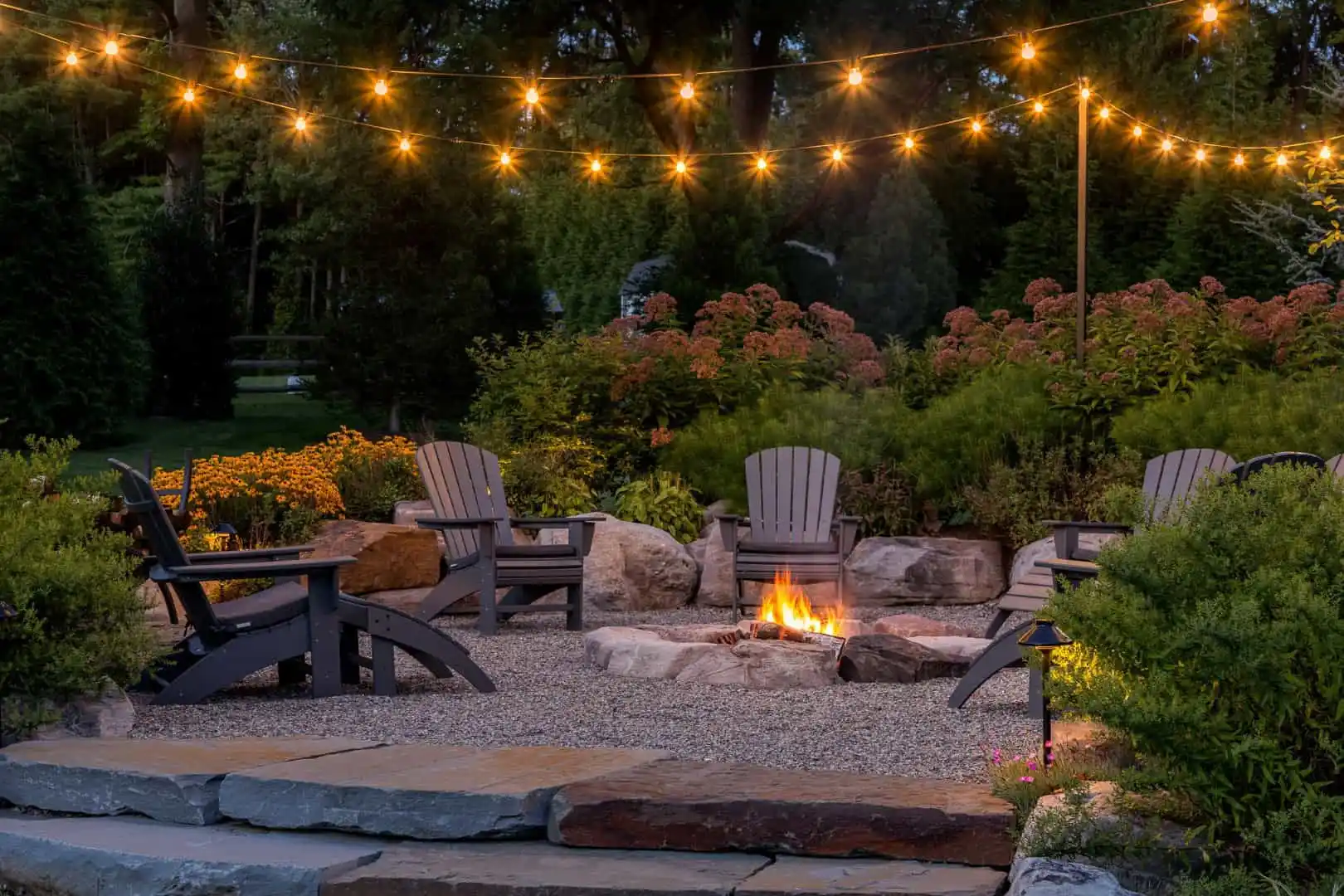 A cozy outdoor patio in Delaware County, PA, features a stone fire pit surrounded by Adirondack chairs. String lights illuminate the garden with blooming flowers and lush greenery, crafted by premier hardscaping services to create a warm and inviting atmosphere.