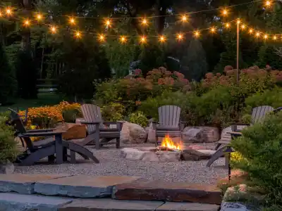 A cozy outdoor patio in Delaware County, PA, features a stone fire pit surrounded by Adirondack chairs. String lights illuminate the garden with blooming flowers and lush greenery, crafted by premier hardscaping services to create a warm and inviting atmosphere.