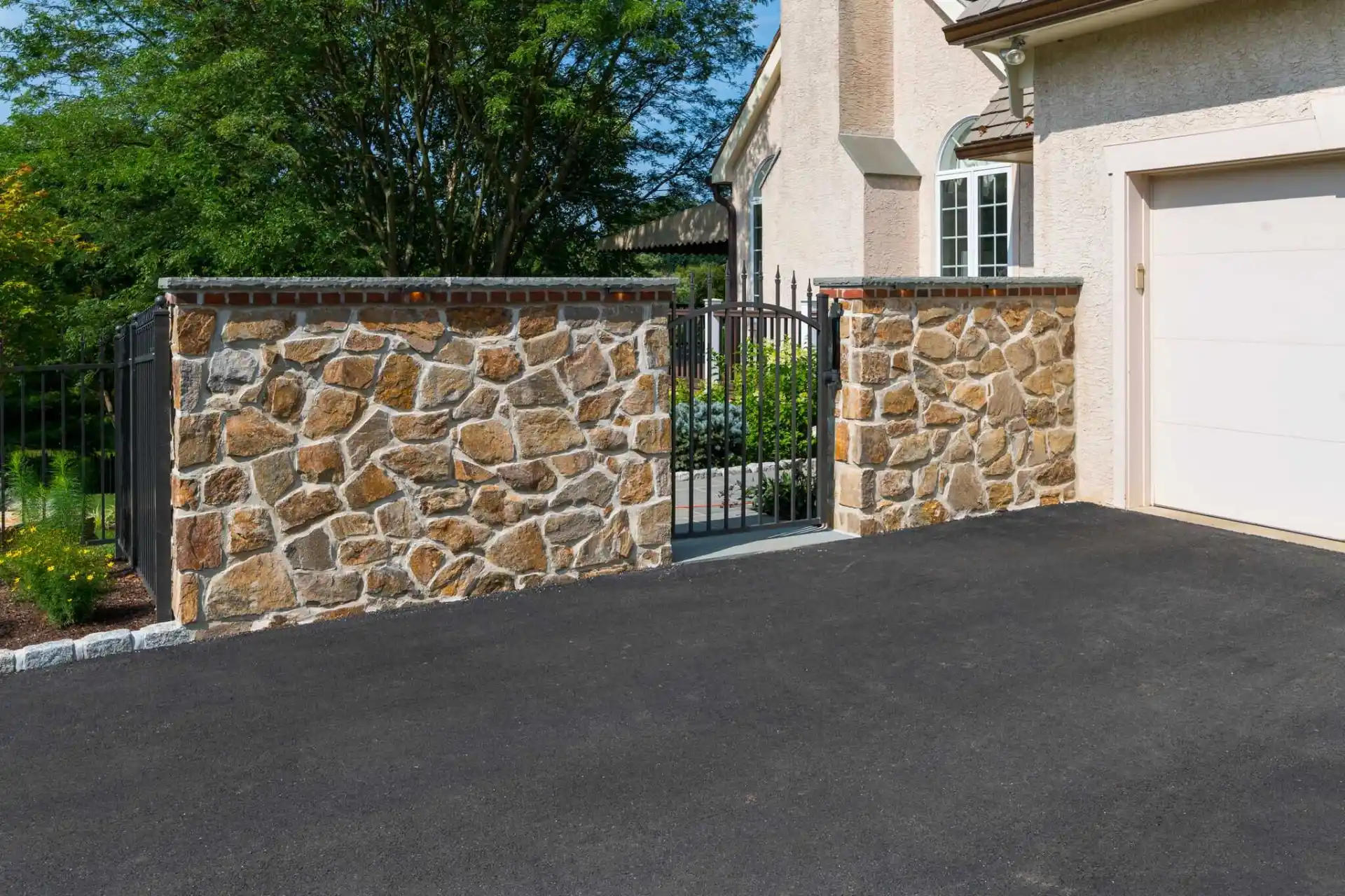 A black metal gate set between two stone pillars opens onto a pathway leading to a house, with an adjacent light-colored garage and lush greenery in the background.
