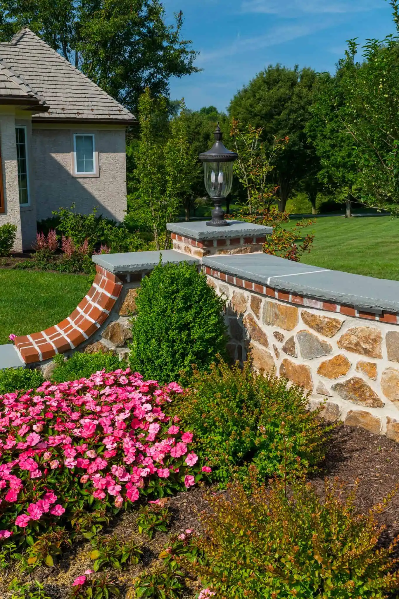 A garden with blooming pink flowers, green shrubs, and a stone wall topped with a lantern, set beside a house with a tiled roof and surrounded by green grass and trees.