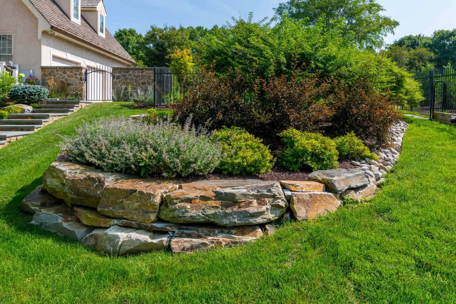 A landscaped garden with layered stone retaining walls, green shrubs, and flowering plants, set in a grassy yard beside a house and fenced area.