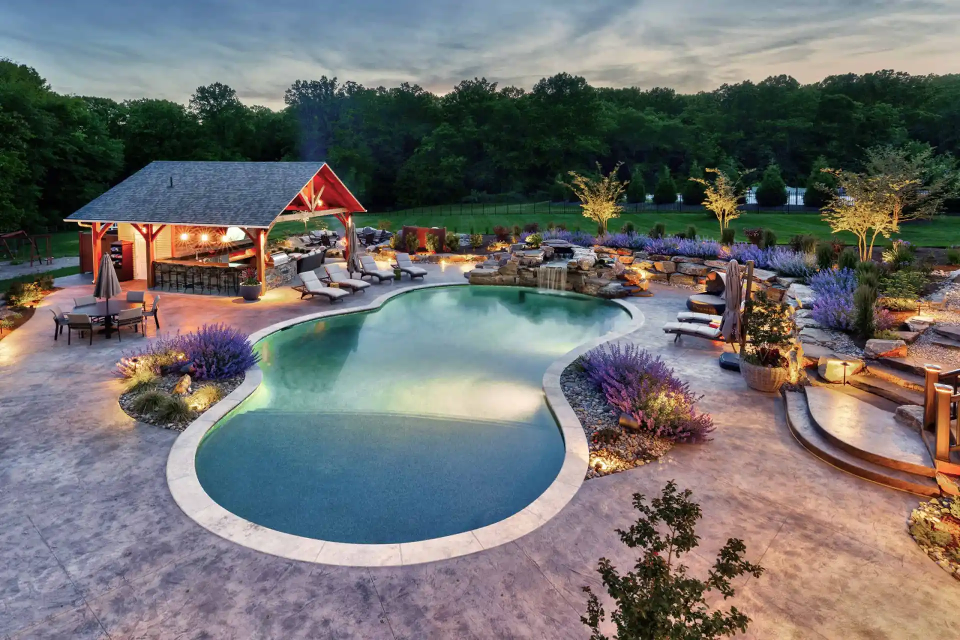 A luxurious backyard pool with curved edges, surrounded by lounge chairs, a covered outdoor bar, landscaped gardens, purple flowers, and soft evening lighting, with trees and grass in the background.
