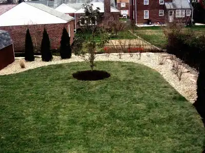 A backyard in Delaware County, PA, showcases outdoor living with a circular grass patch centered by a tree. A border of white pebbles and small bushes surrounds it, while brick houses and a shed are framed by triangular shrubs, offering an idyllic retreat with exquisite landscaping services.