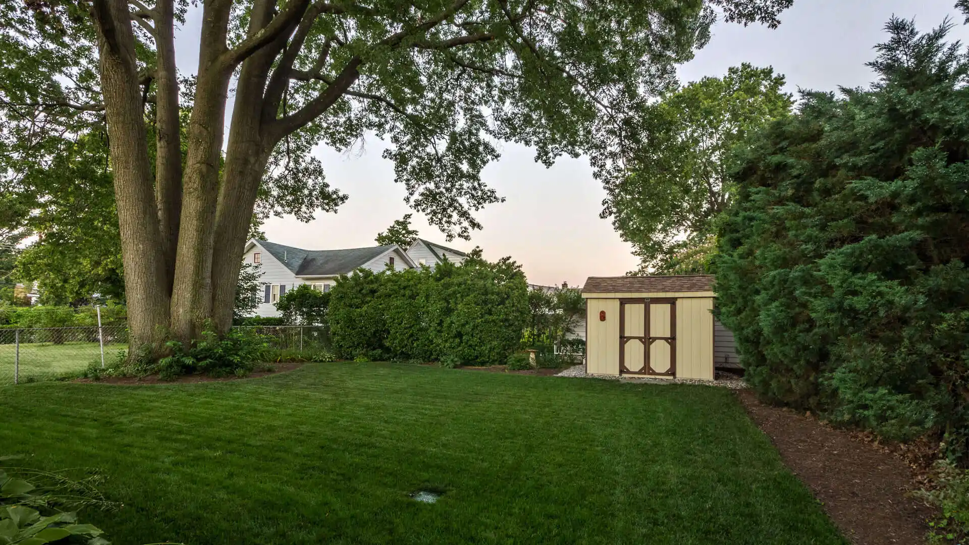 A neatly manicured backyard with lush green grass, large trees providing shade, and a small storage shed near the right corner reflects expert landscaping services in Delaware County, PA. A traditional-style house with white siding is partially visible in the background.