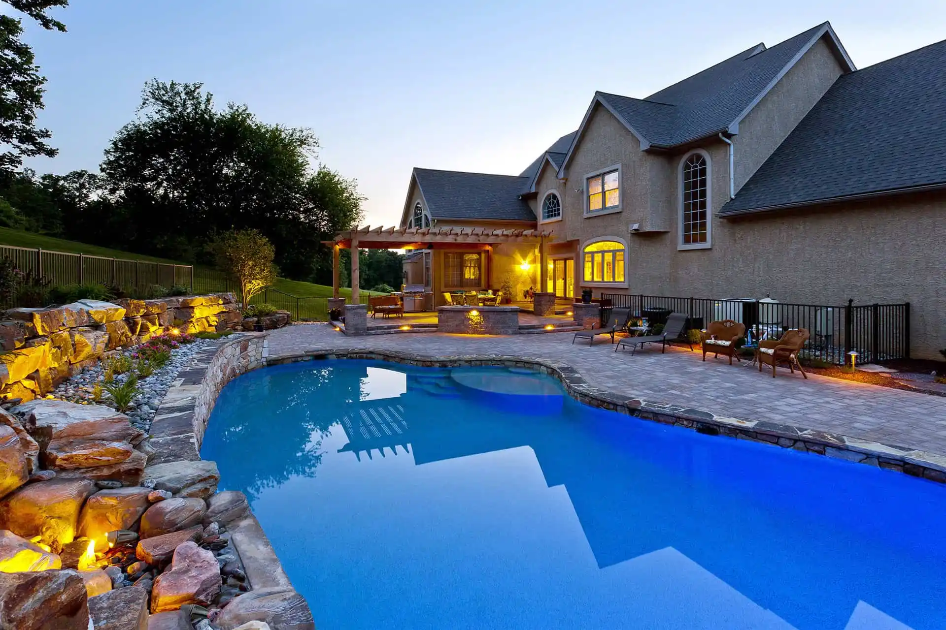 A large house with multiple windows overlooks a backyard featuring a swimming pool, patio with seating, pergola, stone landscaping, outdoor lighting, and lush greenery at dusk.
