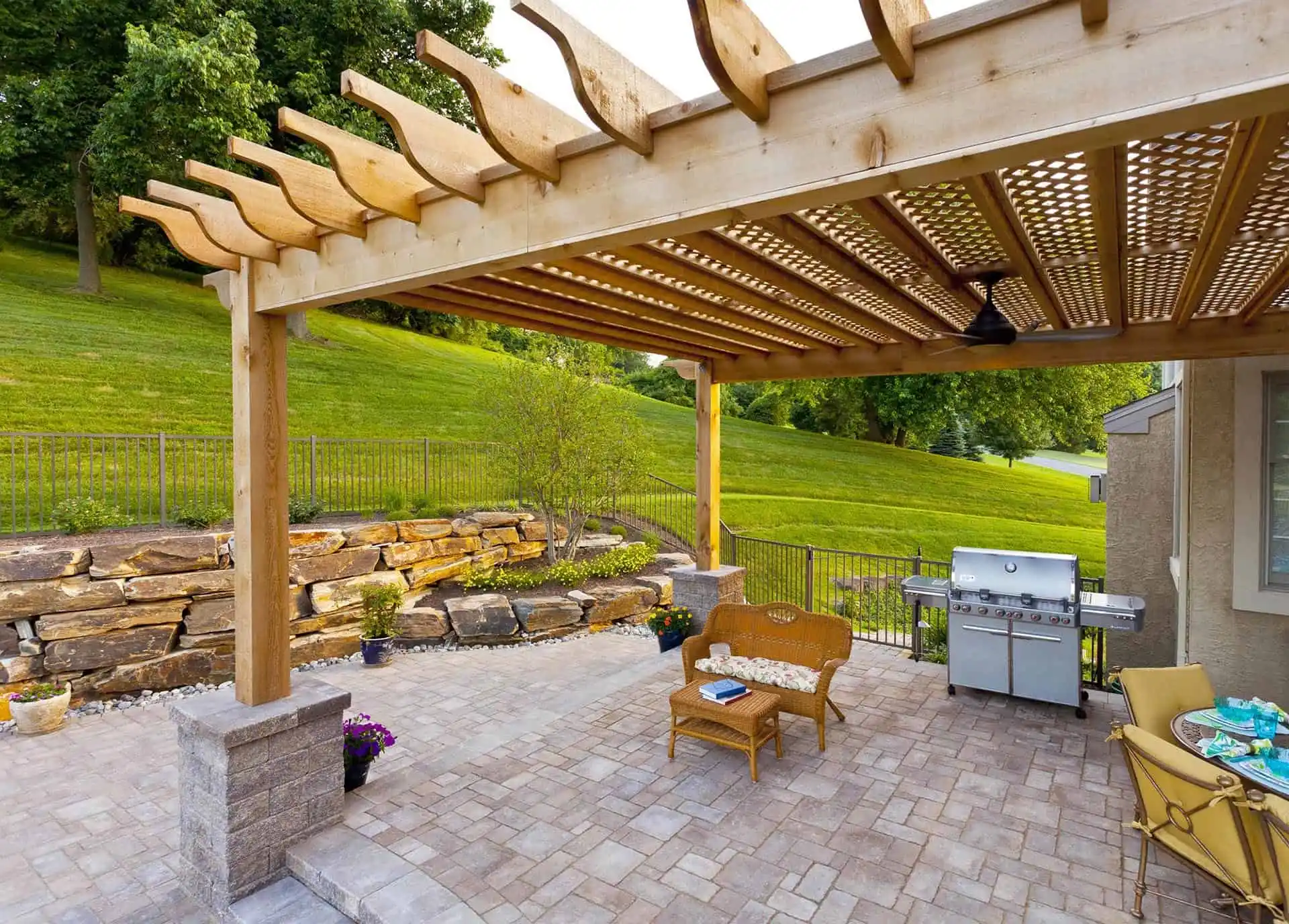 A backyard patio with a wooden pergola, outdoor seating, a small coffee table, a barbecue grill, potted plants, and a lush green hillside with stone retaining walls in the background.