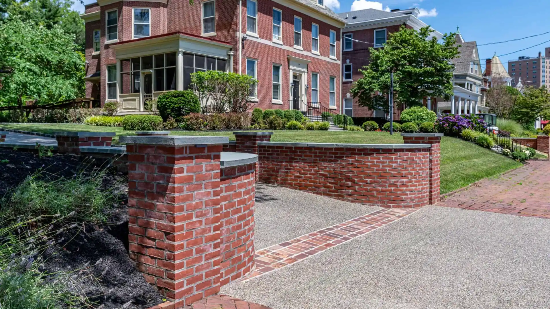 Red brick house with a covered porch and dormer windows, surrounded by a manicured lawn and shrubs. A brick wall frames the driveway, enhanced by expert landscaping services in Delaware County, PA. Above, the clear blue sky hosts a few wispy clouds.