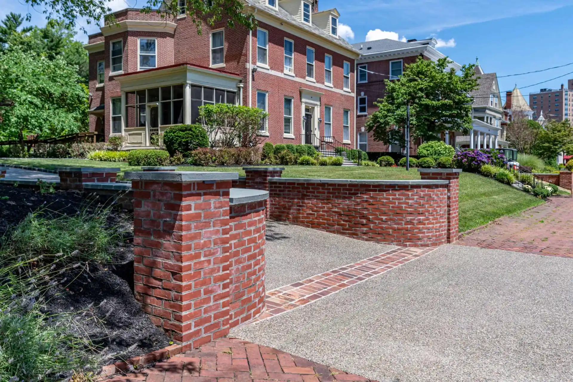 Red brick house with a covered porch and dormer windows, surrounded by a manicured lawn and shrubs. A brick wall frames the driveway, enhanced by expert landscaping services in Delaware County, PA. Above, the clear blue sky hosts a few wispy clouds.