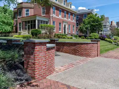 Red brick house with a covered porch and dormer windows, surrounded by a manicured lawn and shrubs. A brick wall frames the driveway, enhanced by expert landscaping services in Delaware County, PA. Above, the clear blue sky hosts a few wispy clouds.