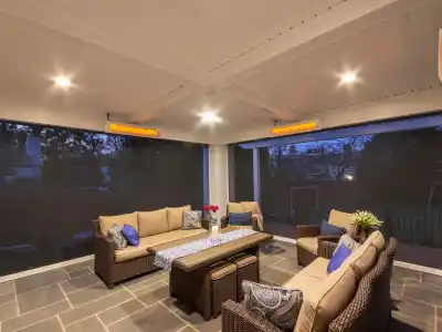 A cozy outdoor patio with wicker furniture, cushioned seats, and blue pillows showcases exquisite outdoor living Delaware County PA style. A table with a blue runner and vase of red flowers is at the center. Overhead heaters are on, and the area is enclosed by screens. Twilight view in the background.