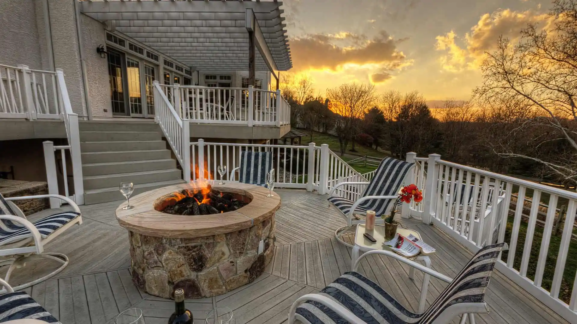 A cozy outdoor deck in Delaware County, PA, showcases a stone fire pit amidst striped lounge chairs and wine glasses. As the sun sets over a scenic landscape with trees, it casts a warm glow on the wooden deck and white railings—a perfect example of exquisite outdoor living.
