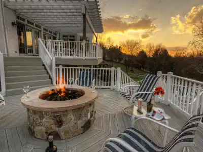 A cozy outdoor deck in Delaware County, PA, showcases a stone fire pit amidst striped lounge chairs and wine glasses. As the sun sets over a scenic landscape with trees, it casts a warm glow on the wooden deck and white railings—a perfect example of exquisite outdoor living.
