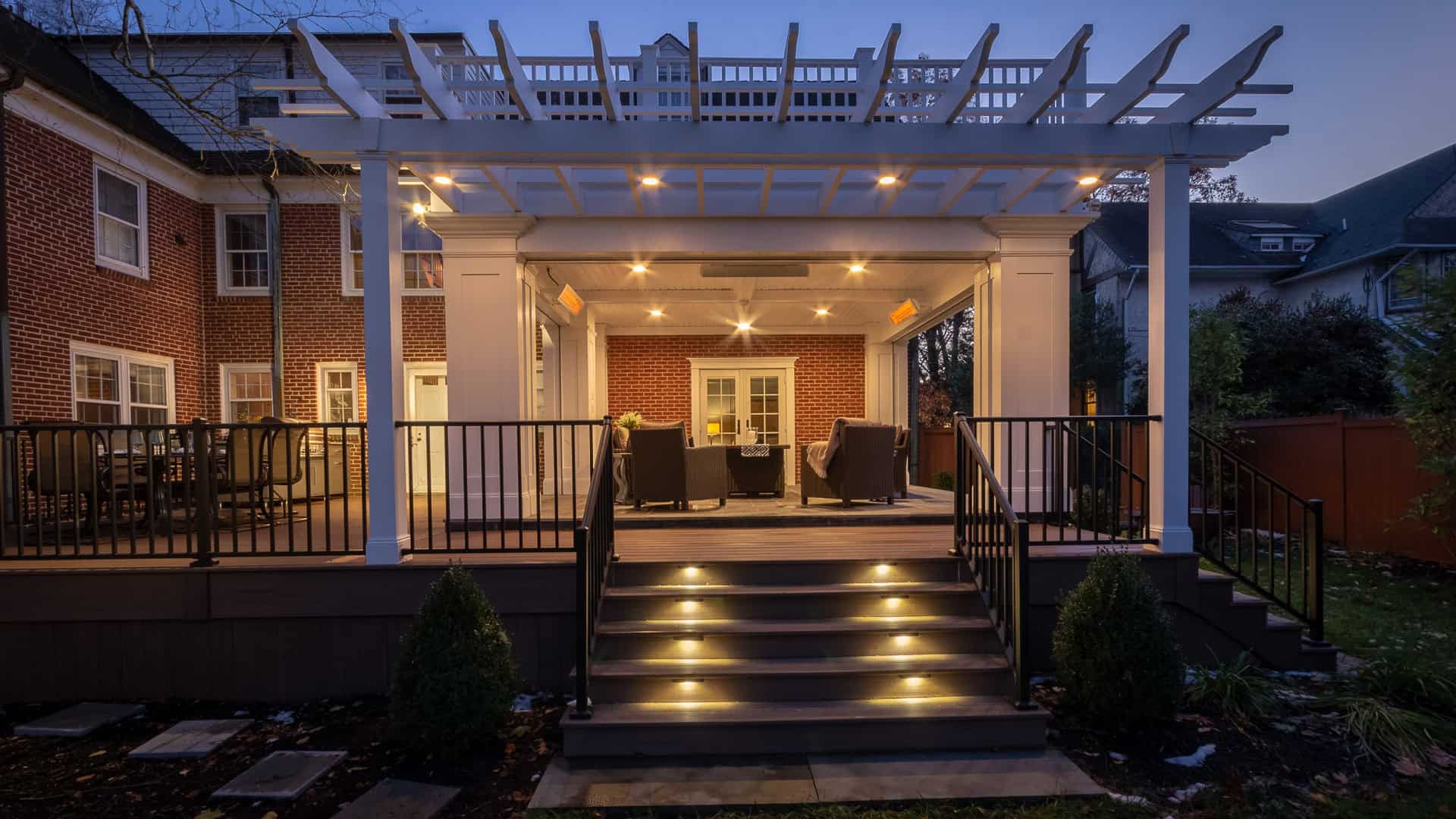 A warmly lit backyard patio with a pergola, steps with built-in lights, black railings, and outdoor seating, attached to a brick house at dusk.