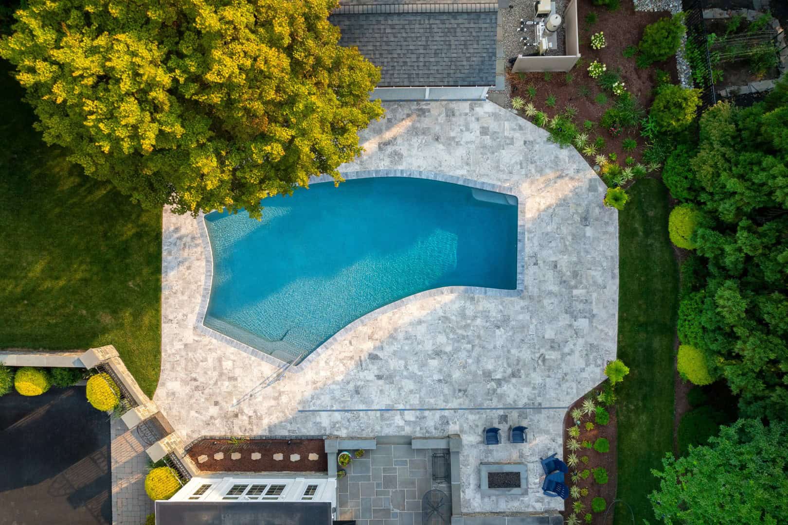 Aerial view of a backyard in Delaware County, PA, featuring a curvy, blue swimming pool surrounded by light stone tiles. Adjacent is a patio with blue chairs. Lush green trees and grass border the area, showcasing expert landscaping services for serene outdoor living.