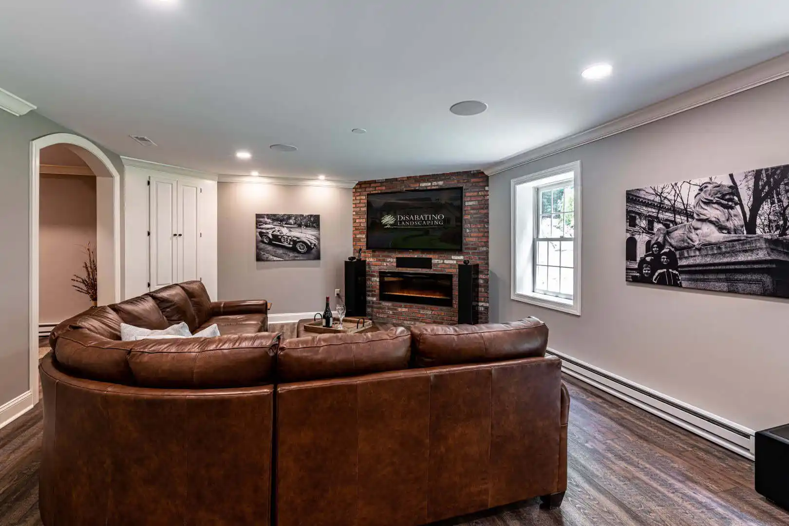 A cozy living room in Delaware County, PA, boasts a brown leather sectional sofa facing a TV on a brick wall above the fireplace. Black-and-white photographs adorn the walls, while natural light floods through the window. A bottle and glasses rest invitingly on the coffee table.