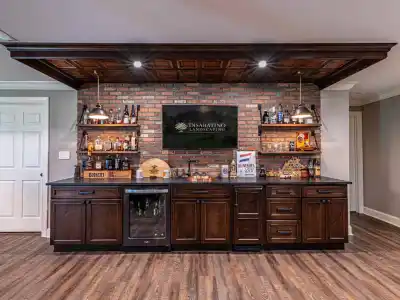 A home bar with a brick accent wall features wooden cabinets and a mini-fridge, seamlessly integrating into the charm of outdoor living. Shelves display liquor bottles and decor. A TV is mounted centrally above the bar, topped with bottles, a sign, and various items.