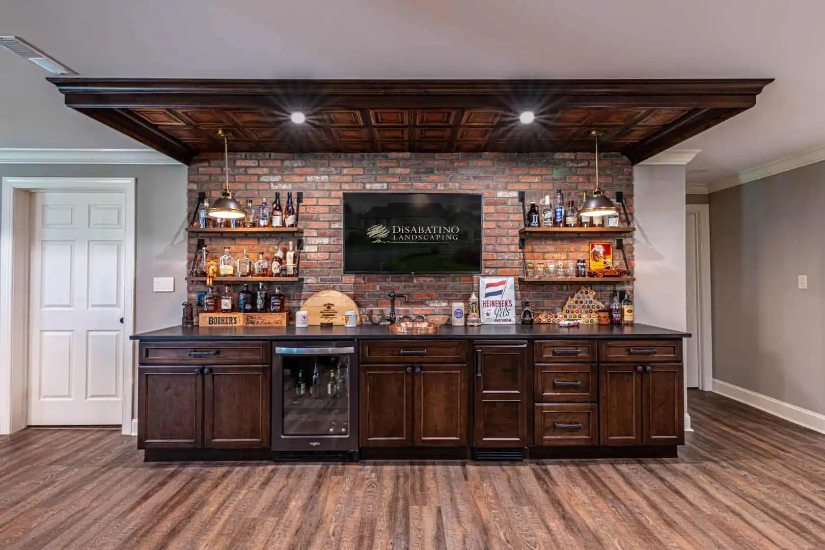 A home bar with dark wood cabinets, a brick backsplash, and liquor shelves is elegantly styled for outdoor living Delaware County PA. A TV showcasing 