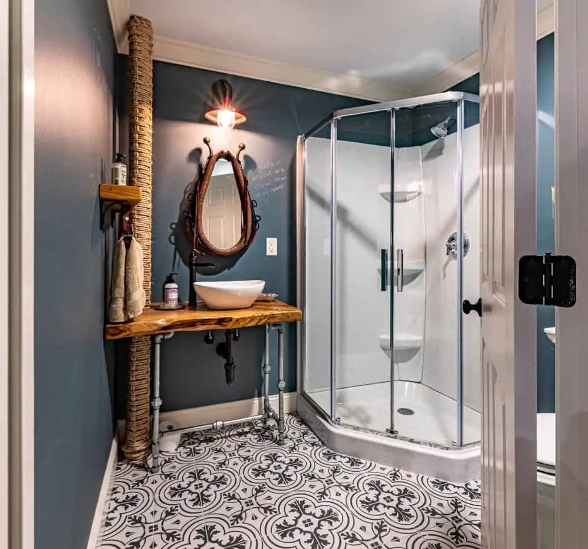 A stylish bathroom with dark blue walls and a patterned tile floor. It boasts a round mirror above a rustic wooden vanity with vessel sink. A glass shower enclosure stands to the right, completing this serene indoor space—a perfect retreat from your outdoor living in Delaware County, PA.