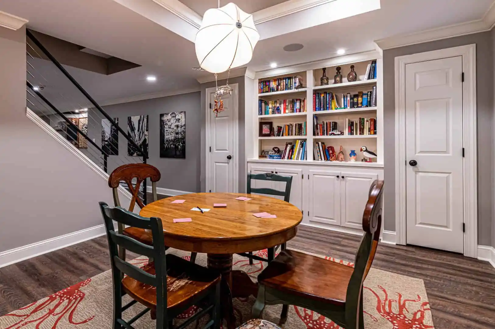 A cozy basement room with a round wooden table and four chairs, perfect for discussing outdoor living in Delaware County, PA. A bookshelf filled with books and decor stands against the wall. A modern pendant light hangs above, while a staircase with cable railing is visible over the patterned rug.