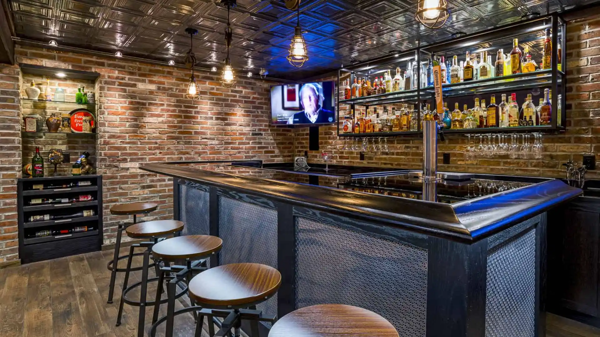 Stylish home bar with a black counter and four stools. Shelves stocked with bottles, a TV displaying news, brick walls, and ambient lighting create a cozy atmosphere—perfect for unwinding after enjoying your outdoor living space in Delaware County, PA.