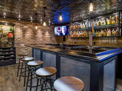 Stylish home bar with a black counter and four stools. Shelves stocked with bottles, a TV displaying news, brick walls, and ambient lighting create a cozy atmosphere—perfect for unwinding after enjoying your outdoor living space in Delaware County, PA.