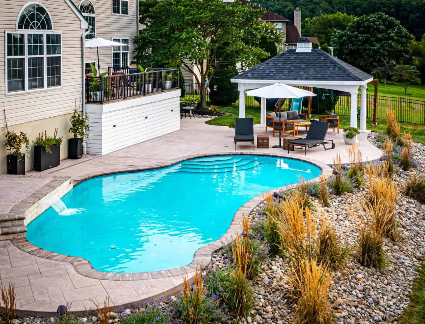 A backyard oasis offers a kidney-shaped swimming pool with clear blue water, framed by beige stone tiles. Discover outdoor living in Delaware County, PA, under a wooden gazebo with a white canopy. Lounge chairs and elegant decorative grasses complete this serene retreat.